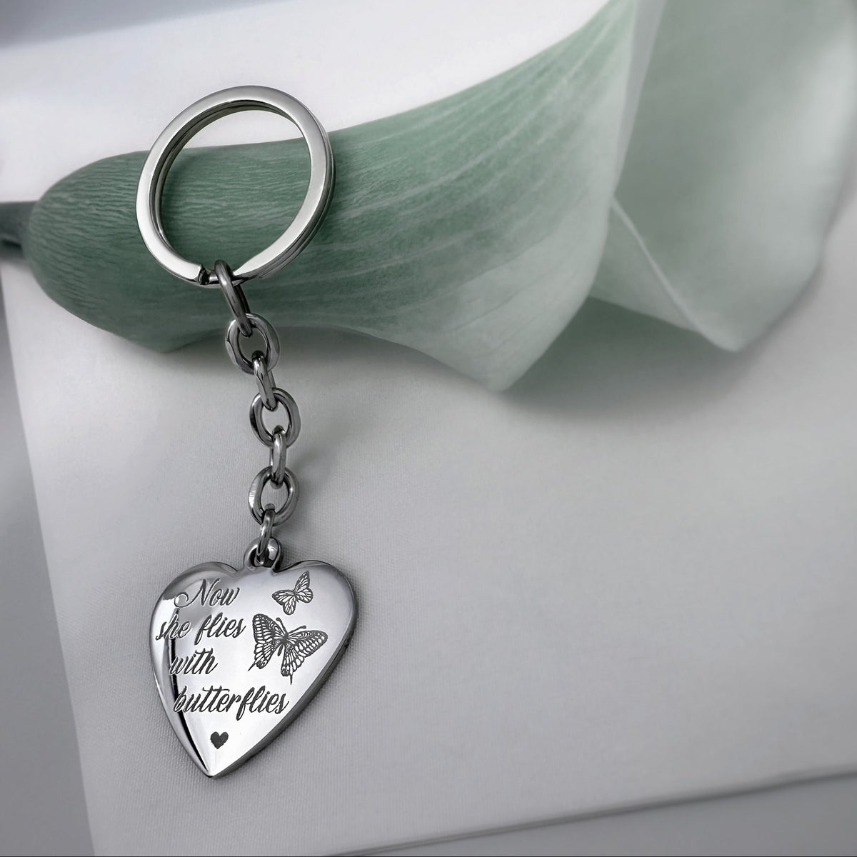 A silver keychain with a heart-shaped charm engraved with customizable message 'Now she flies with butterflies' and butterfly design, resting on a white surface next to a pale green flower.