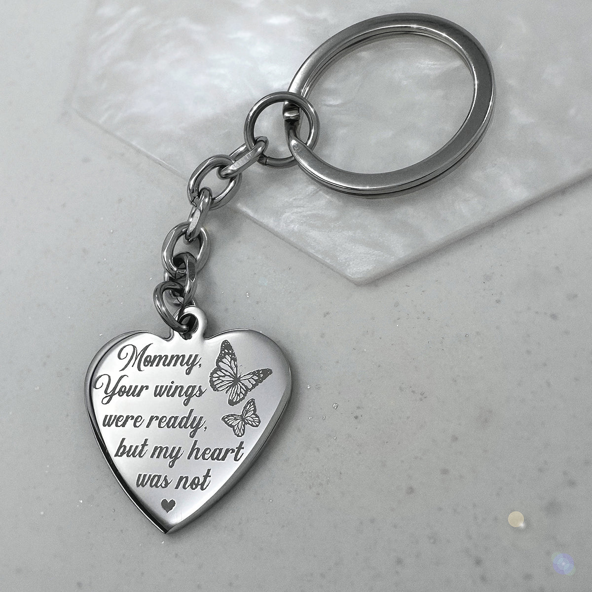 A silver heart-shaped keychain engraved with the custom message 'Mommy, your wings were ready, but my heart was not' alongside butterfly design, shown on a light gray textured surface.