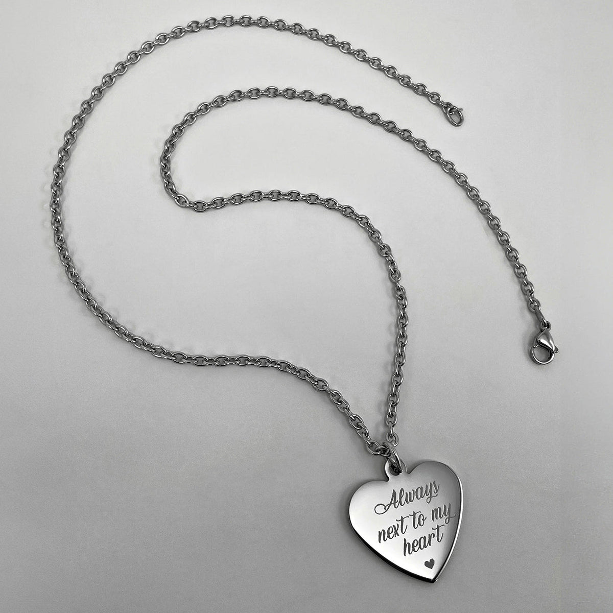 A silver heart-shaped pendant necklace engraved with the personalized words 'Always next to my heart' and a small heart, displayed on a light gray background.