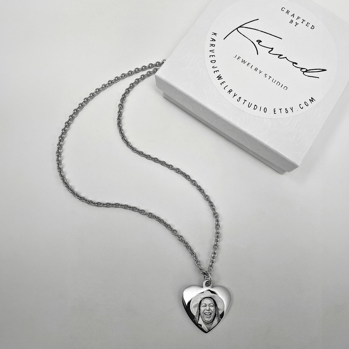 A silver heart-shaped pendant necklace with a custom engraved portrait, displayed next to its white branded Karved Jewelry Studio gift box on a light gray background.