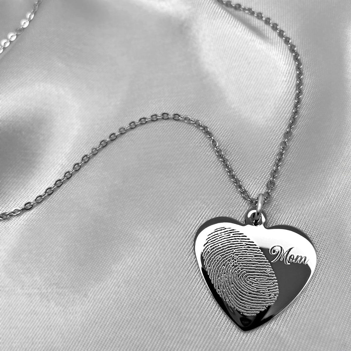 A silver heart-shaped pendant necklace with a custom fingerprint design and the engraved word 'Mom', displayed on draped white satin fabric.