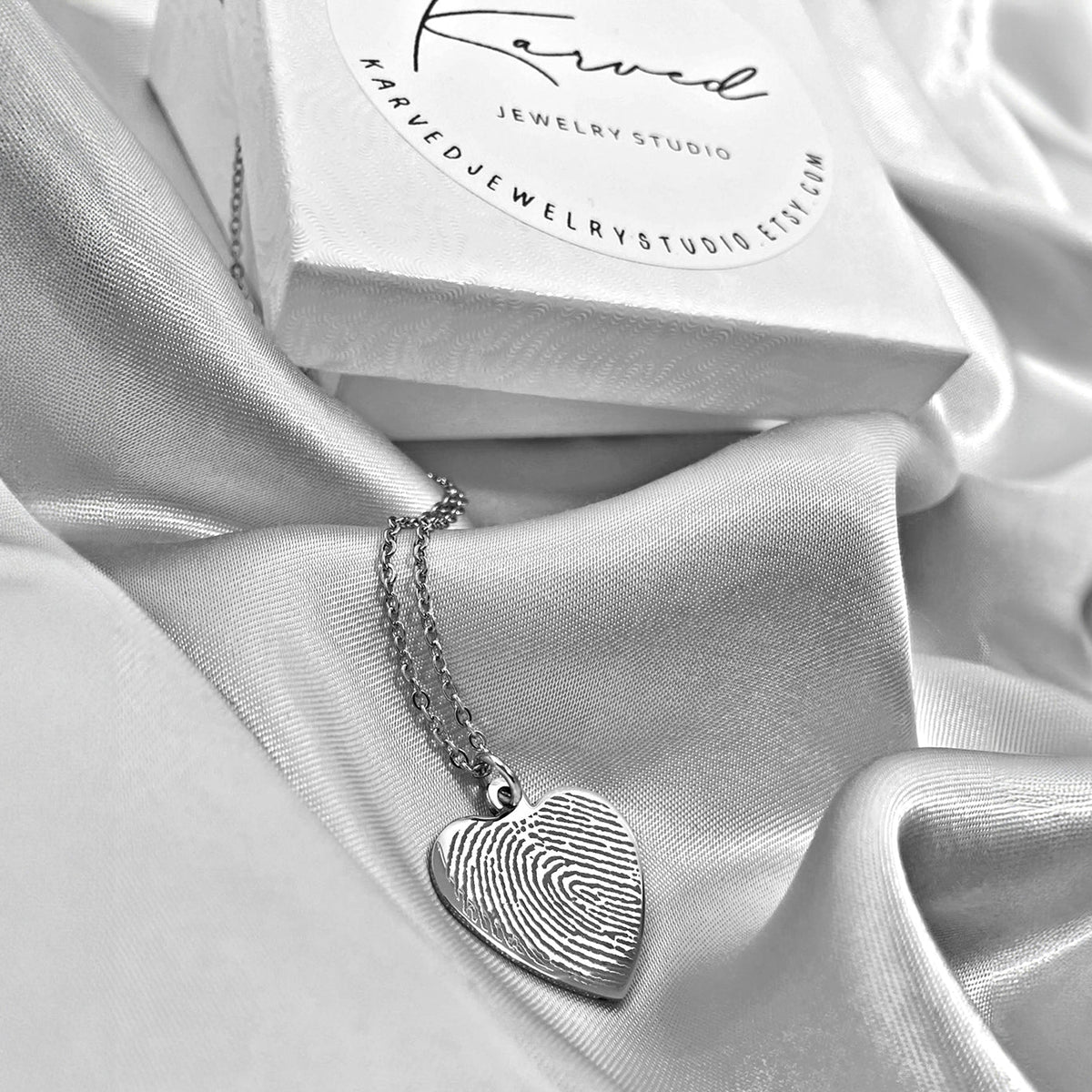A silver heart-shaped pendant with an actual engraved fingerprint, resting on draped white satin fabric next to jewelry box. 