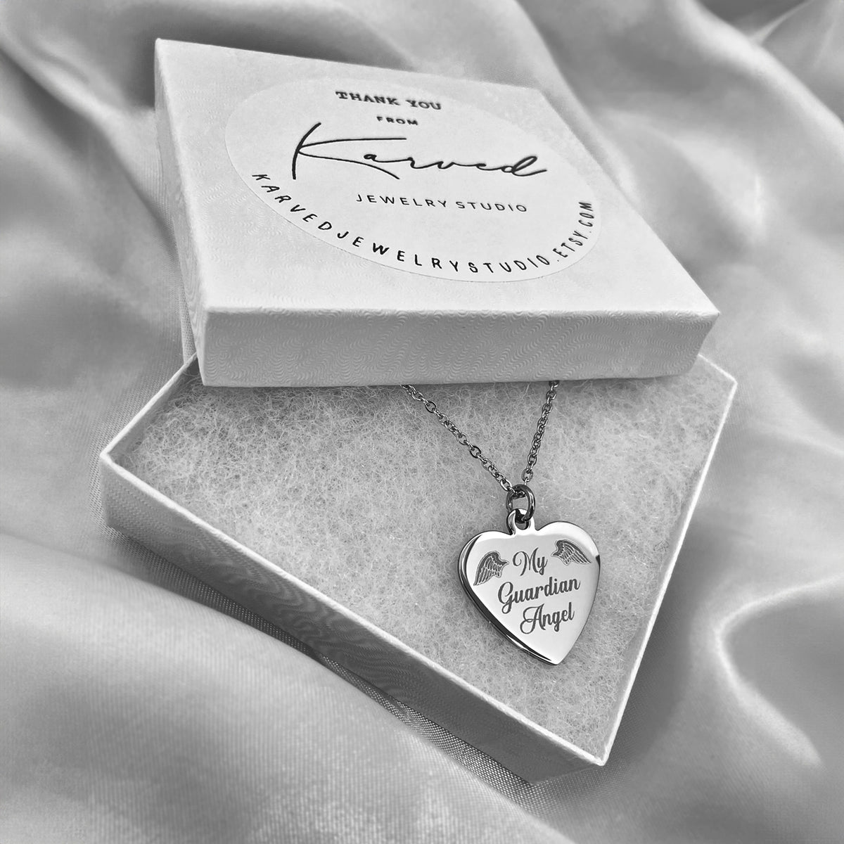 A silver heart-shaped pendant necklace engraved with 'My Guardian Angel' and small angel wings designs, displayed in an open jewelry box