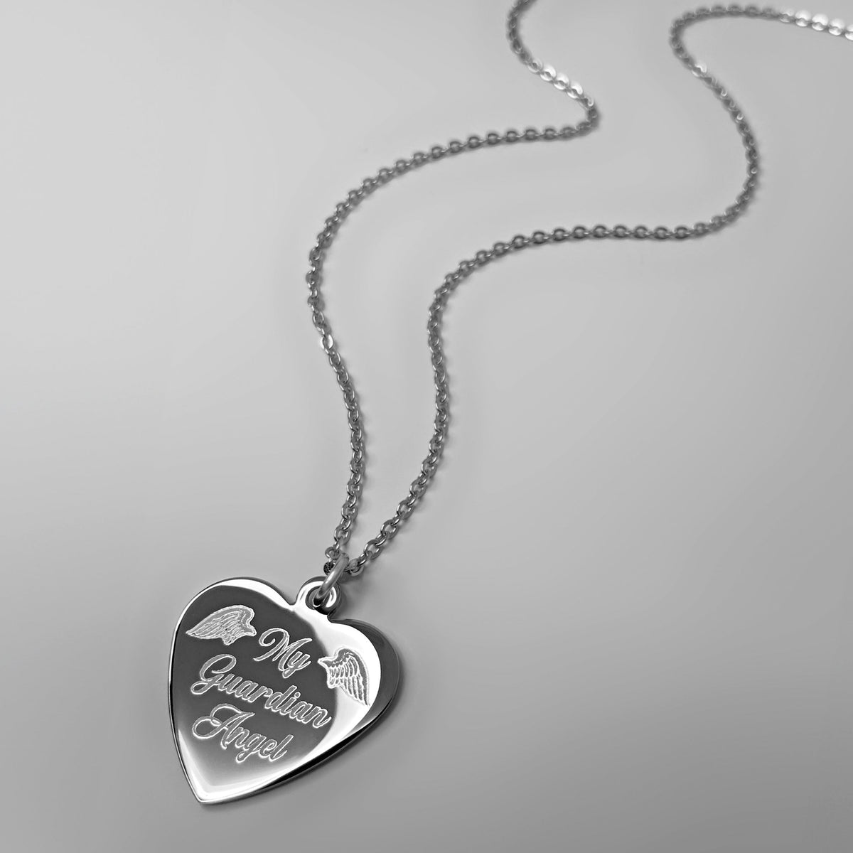 A silver heart-shaped pendant necklace engraved with custom saying 'My Guardian Angel' and small angel wings, displayed on a light gray background.