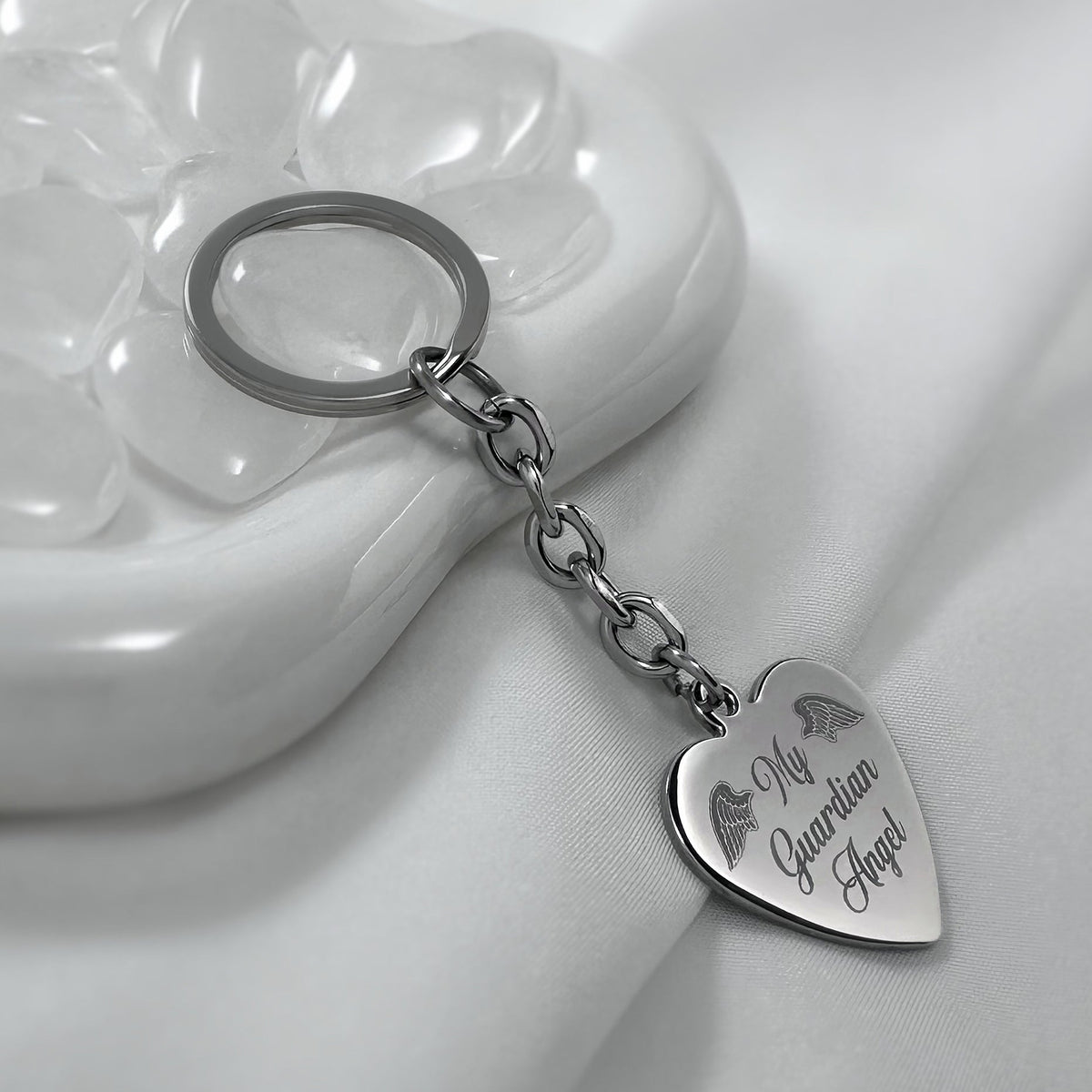 A silver heart-shaped keychain with 'My Guardian Angel' custom engraving, resting on a white ceramic dish with clear stones against a white fabric background.
