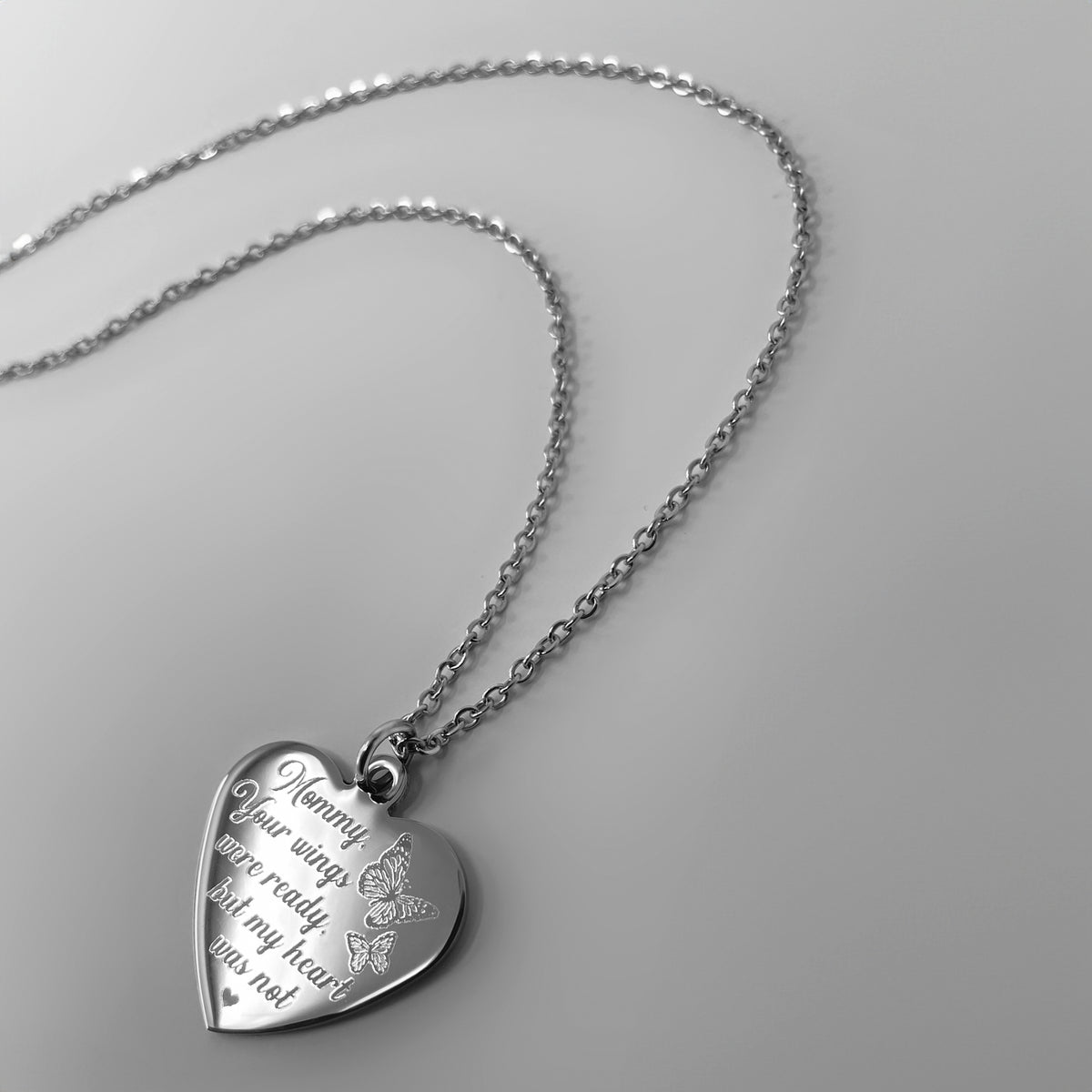 A silver heart-shaped pendant necklace engraved with 'Mommy, your wings were ready, but my heart was not' and a small butterfly design, shown on a light gray background.
