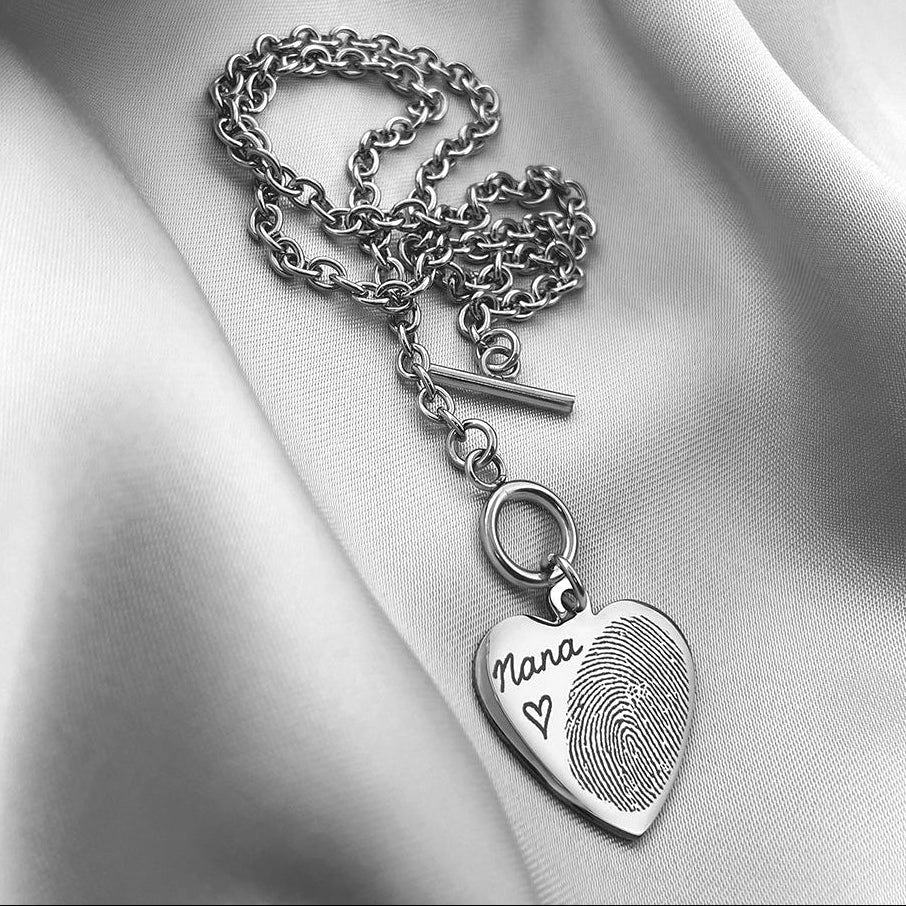 A silver toggle clasp heart necklace engraved with an actual fingerprint and custom handwriting, resting on draped white satin fabric.