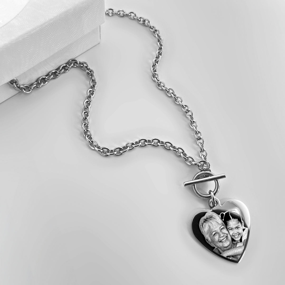 A silver toggle clasp necklace with a heart-shaped pendant featuring a personalized memorial photo of a grandparent, displayed with a gift box on a light gray background.