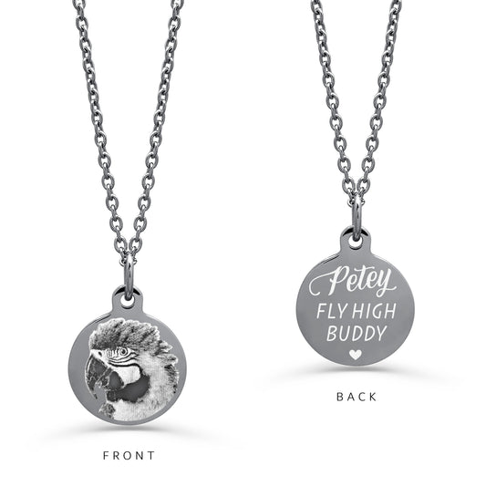 A silver round pendant necklace on a white background. The left side shows a detailed personalized parrot portrait engraved on the pendant, and the right side shows a custom engraved name with a memorial quote.