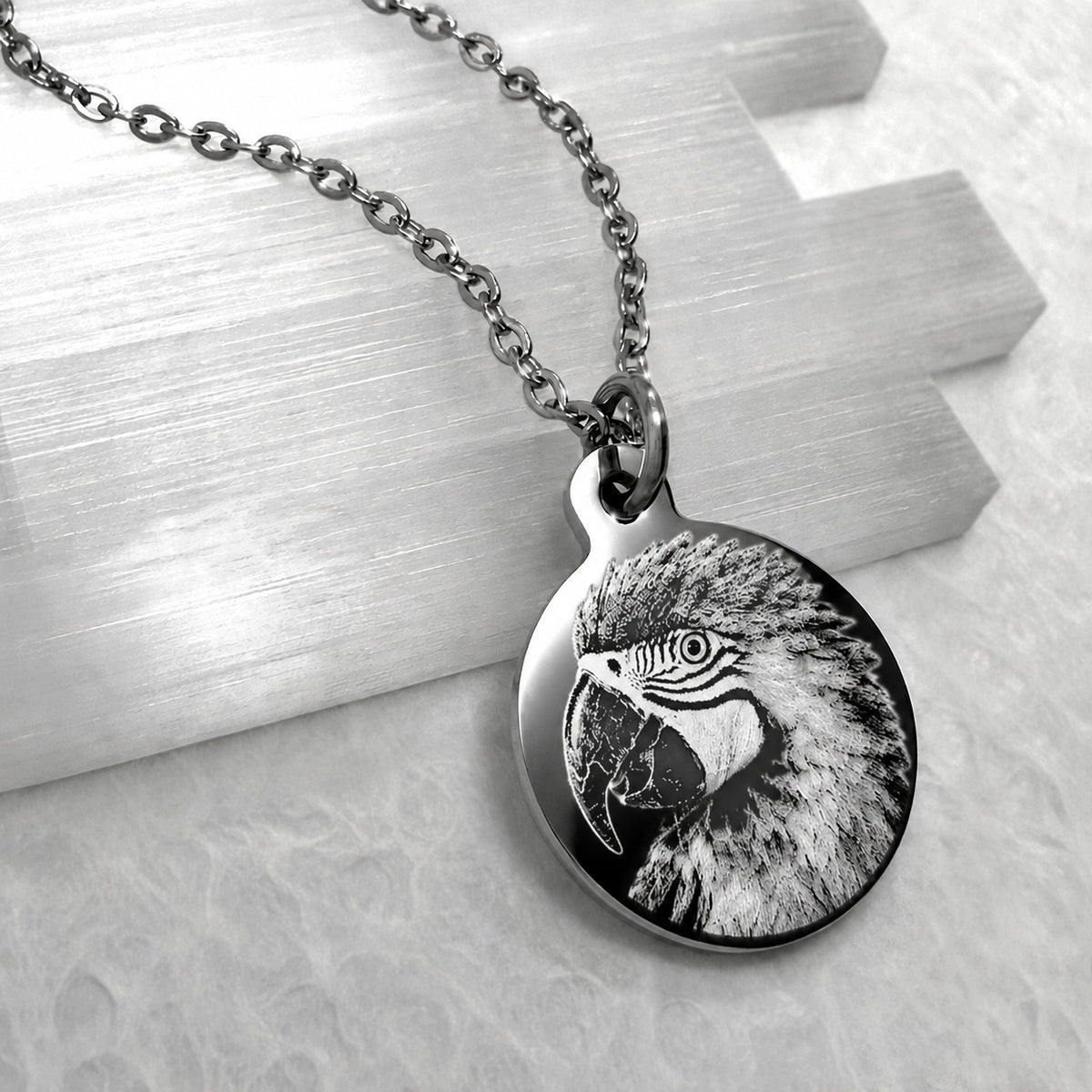 A silver round pendant necklace featuring a detailed engraved pet bird parrot design, with a matching stainless-steel chain on a light textured surface.
