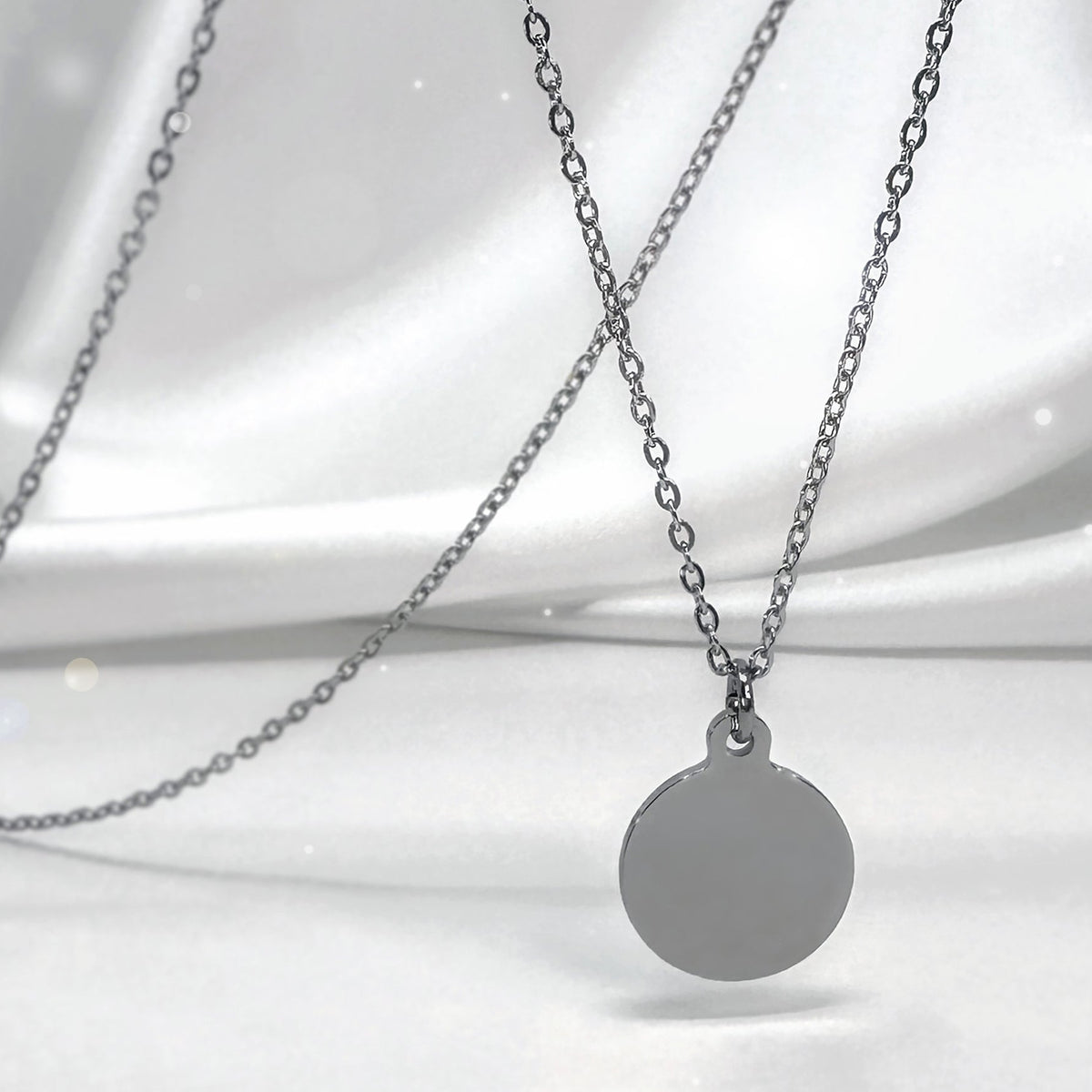 A silver necklace with a round disc pendant, displayed against a draped white satin fabric background.