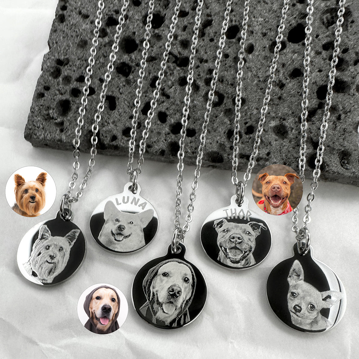 Six silver chain necklaces with round pendants displayed on a white surface next to a dark stone slab. Each pendant features a custom pet portrait, including personalized portraits and photos of different dog breeds.