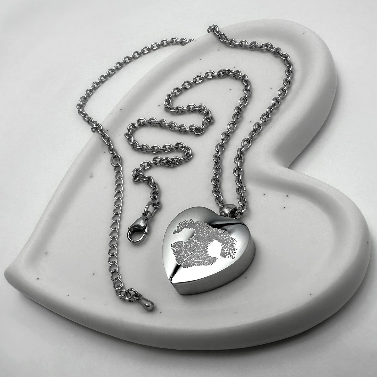 A stainless-steel heart-shaped urn pendant necklace with a personalized paw print engraving, displayed on a white ceramic heart-shaped dish against a light gray background.
