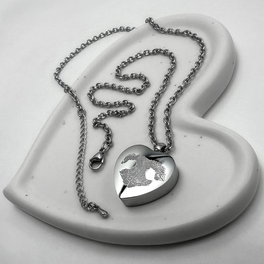 A stainless-steel heart-shaped urn pendant necklace with a personalized paw print engraving, displayed on a white ceramic heart-shaped dish against a light gray background.