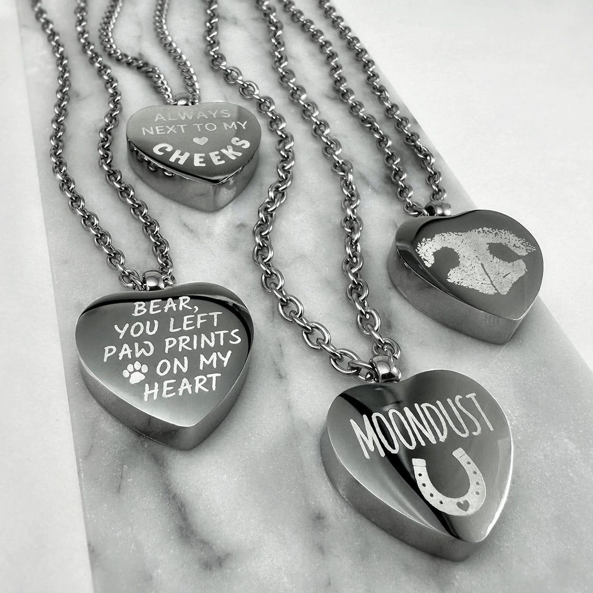 Four heart-shaped urn pendant necklaces with stainless steel chains arranged on a light marble surface. The pendants are engraved with personalized pet memories: one says 'BEAR, YOU LEFT PAW PRINTS ON MY HEART' with a paw print, one says 'ALWAYS NEXT TO MY CHEERS,' one shows a paw and heart outline, and one says 'MOONDUST' with a horseshoe.