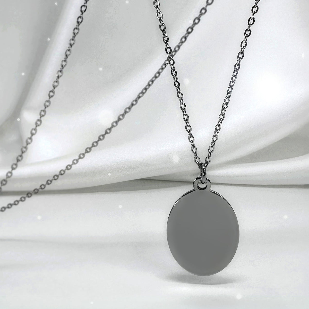 A silver oval pendant necklace with a matching chain, displayed against a draped white satin fabric background.