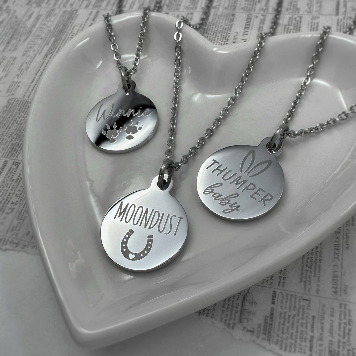 Three silver round pendant necklaces displayed on a white ceramic heart-shaped dish. The pendants are engraved with the custom pet names and actual paw prints: 'Winnie' with a paw print, 'MOONDUST' with a horseshoe, and 'THUMPER baby' with a bunny outline.