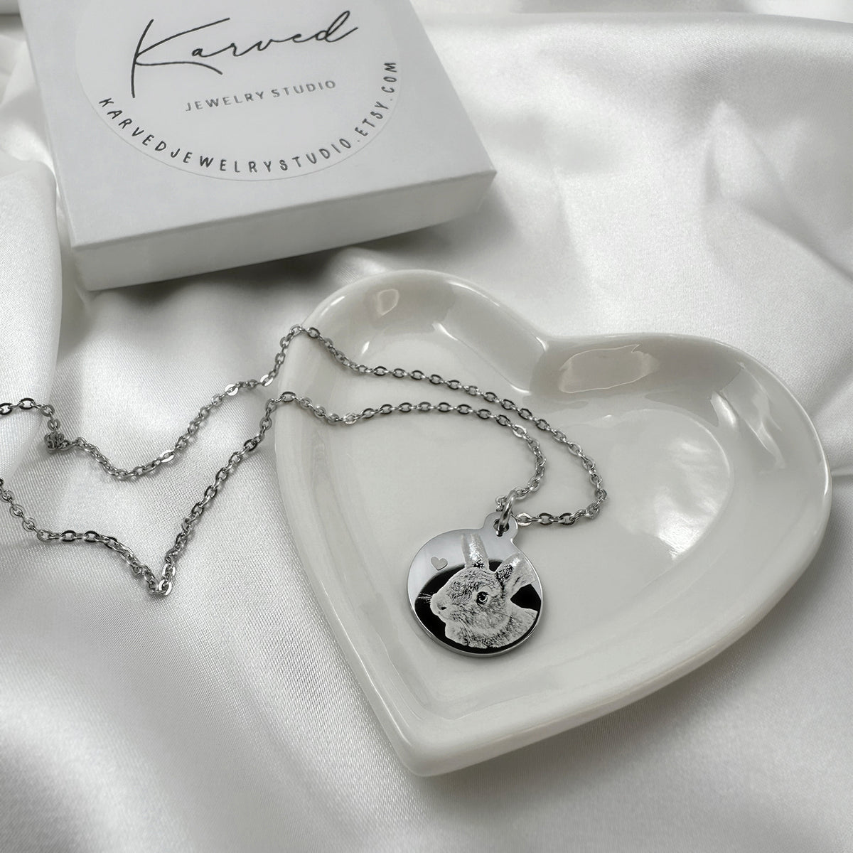 A silver necklace with a round pendant featuring a personalized cat picture, displayed on a white heart-shaped tray next to a KJS jewelry box on a white satin background.