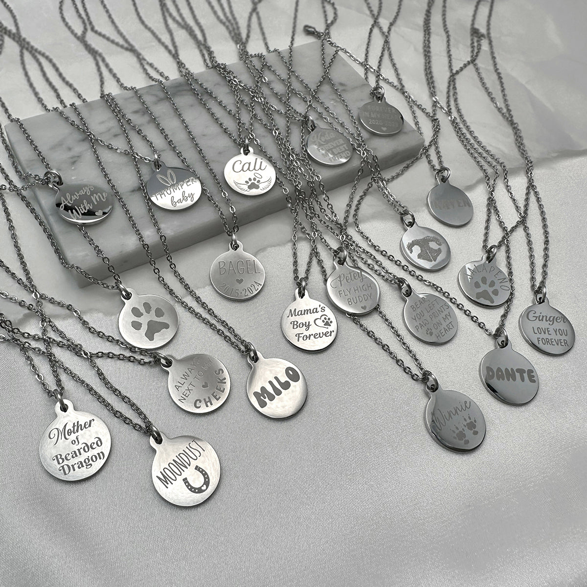 A collection of silver round custom pendant necklaces with engraved personalized names, paw prints, and text, arranged on a light gray fabric surface with a marble block prop.