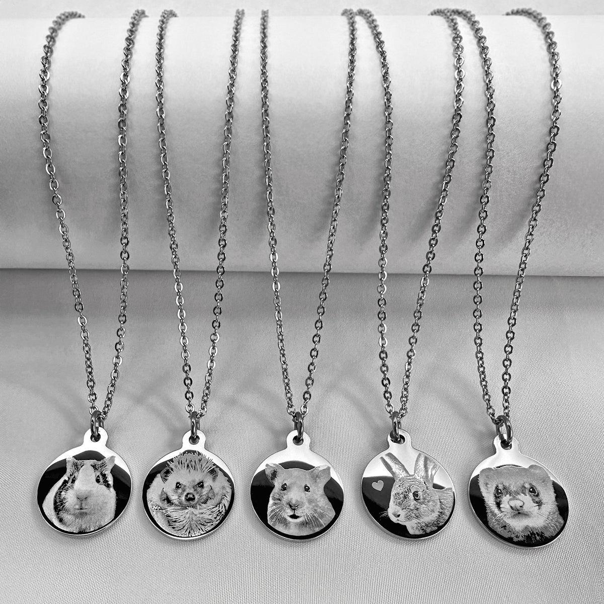 Five silver chain necklaces with round pendants featuring personalized pet portraits, displayed on a white cylindrical stand against a light gray background. 