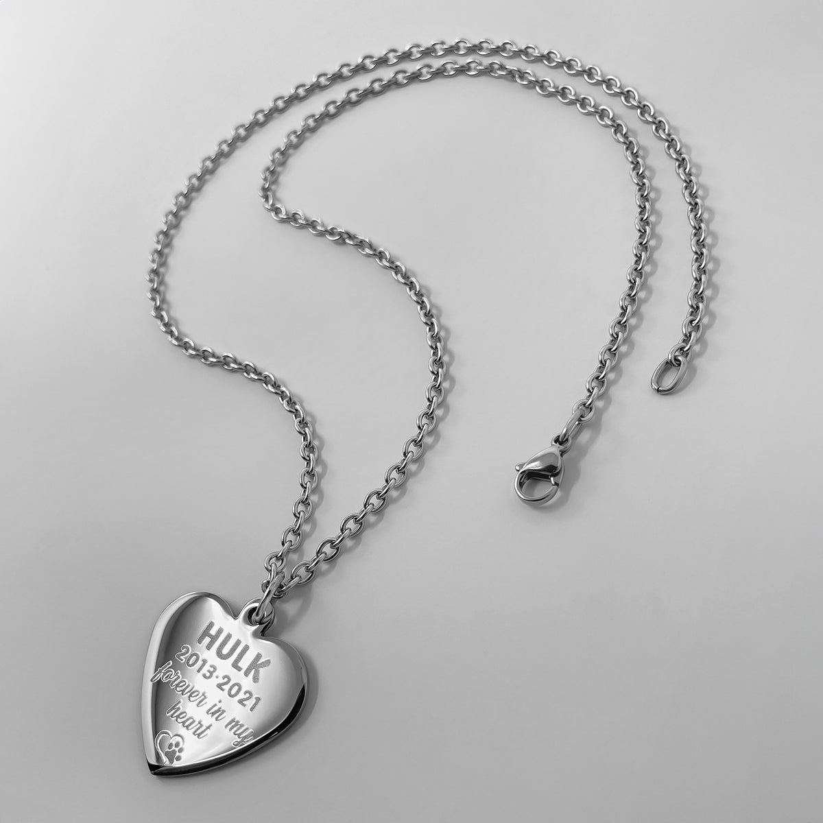 A silver heart-shaped pendant necklace engraved with personalized pet name, custom dates, actual paw print, and personalized memorial message