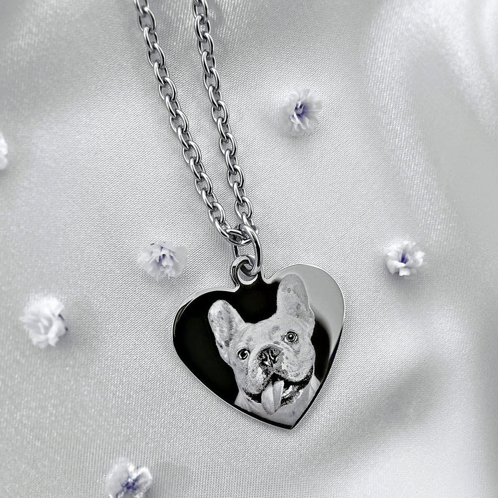 A silver heart-shaped pendant necklace featuring a custom pet portrait of a French Bulldog, displayed on a white satin fabric backdrop.