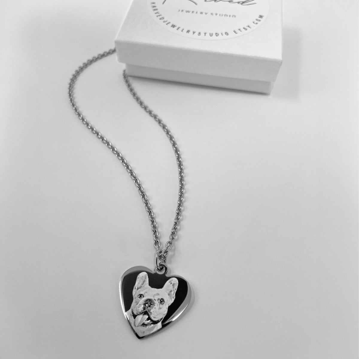 A silver heart-shaped pendant necklace featuring a custom engraved pet portrait, displayed on a light gray surface next to its white jewelry box.