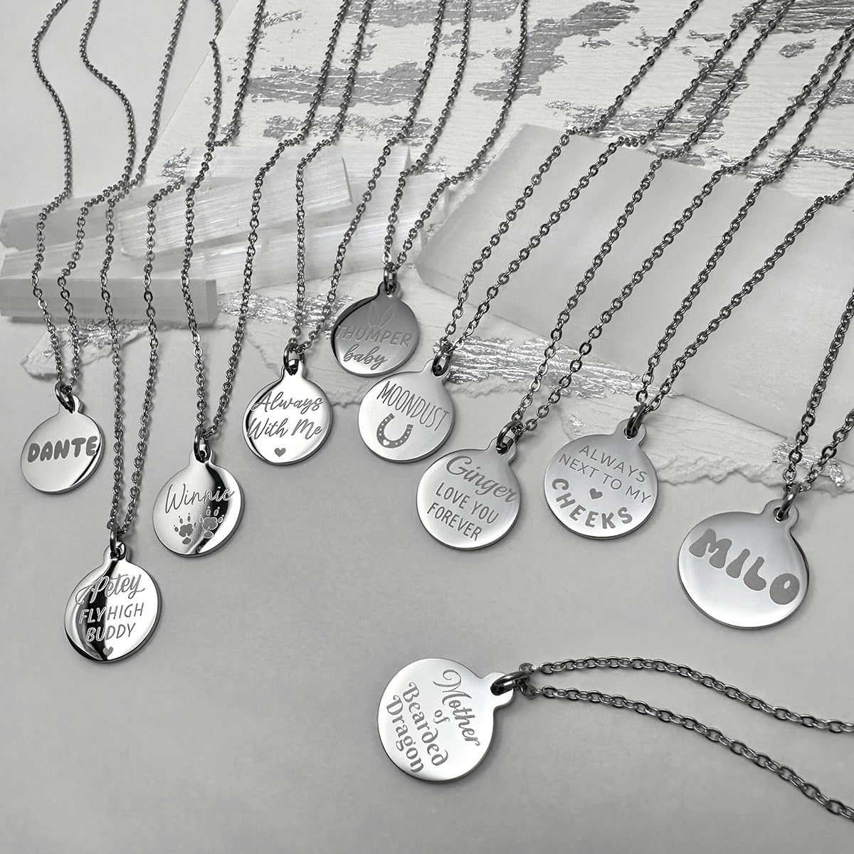 A set of personalized silver round pendant necklaces with custom engraved names and personalized messages, arranged on a light gray surface with a white textured background.