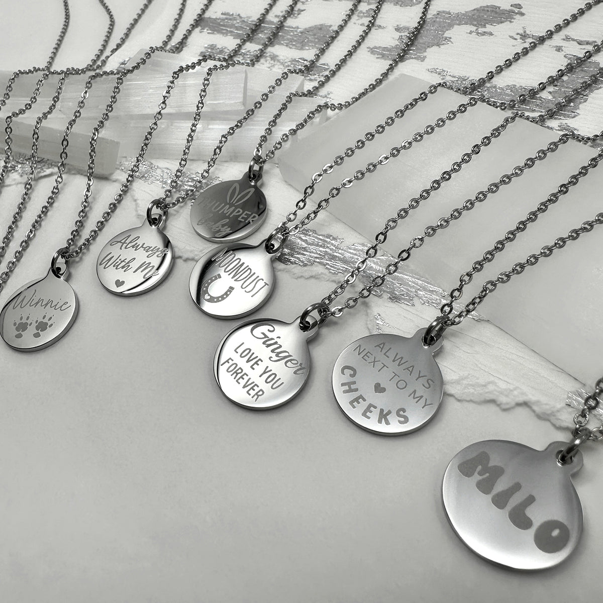 A set of silver round personalized pendant necklaces with engraved names and designs.