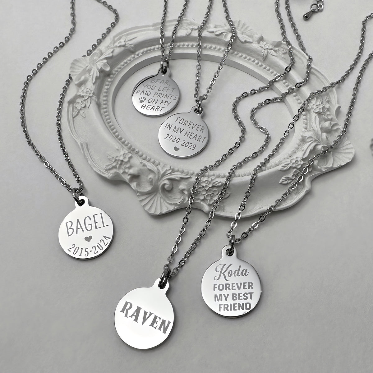 Five silver round pendant necklaces with engraved personalized names and dates are arranged on a white decorative tray against a light gray background. The pendants include 'BAGEL 2015-2024', 'RAVEN', 'Koda FOREVER MY BEST FRIEND', 'FOREVER IN MY HEART 2021-2023', and 'YOU ARE MY HEART'.