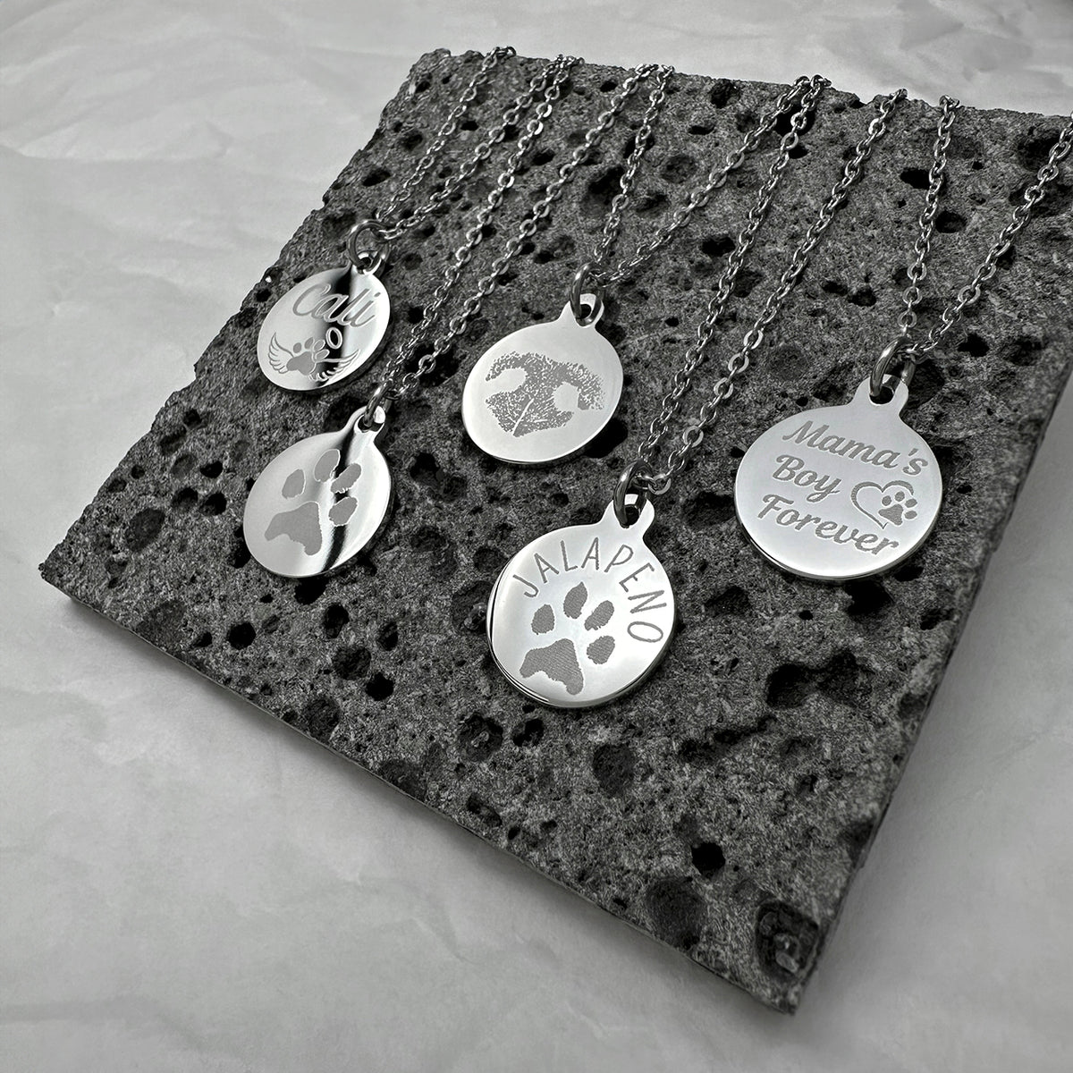 Five silver pet personalized and memorial necklaces with round engraved pendants. The pendants feature designs including paw prints, a heart with a dog portrait, custom name, actual nose print.