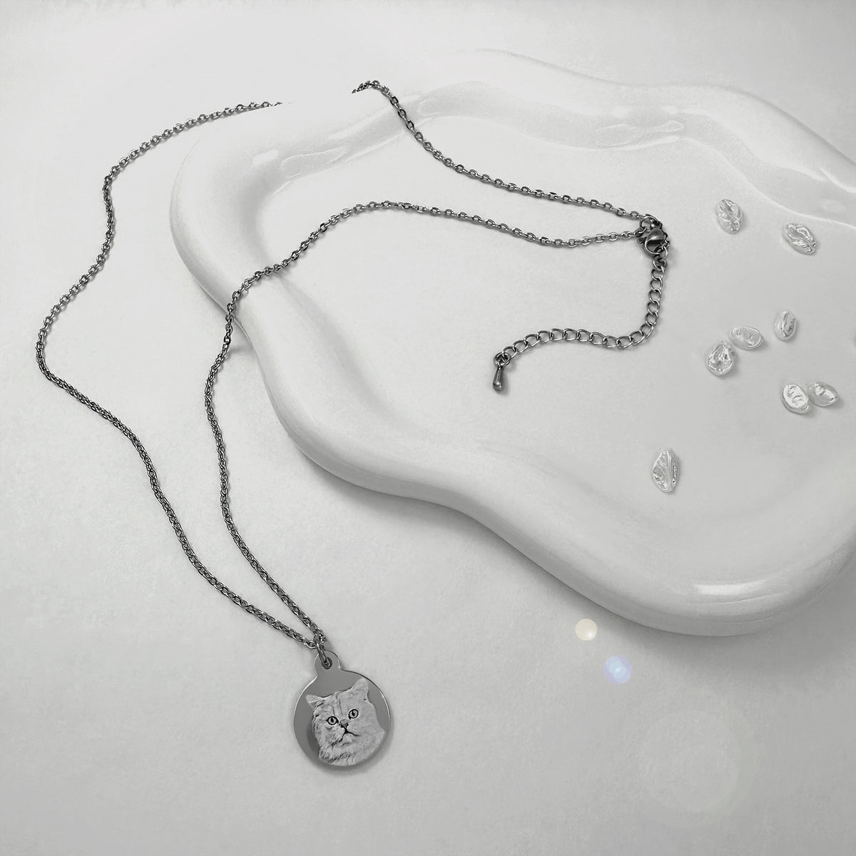 A silver chain necklace with a round pendant engraved with a personalized cat portrait, displayed on a white wavy ceramic dish.