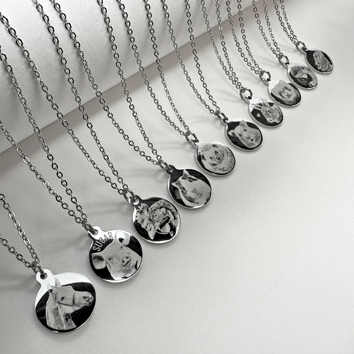 A set of personalized silver necklaces with round custom engraved pendants showing various pet portrait photos, displayed on a light gray surface.